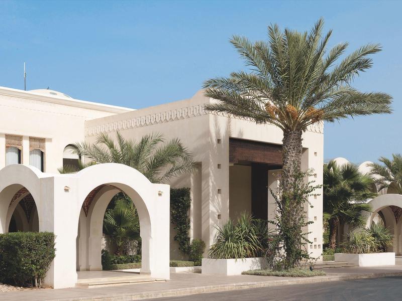 Ingresso dell'hotel con archi bianchi e palme sotto un cielo limpido.
