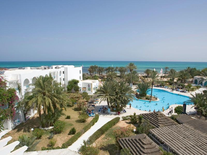 Hotel complex with pool, palm trees, and sea view under clear sky.