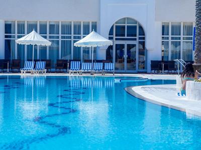 Hotelpool mit blauer Wasseroberfläche, Sonnenliegen und weißen Sonnenschirmen neben einem weißen Gebäude.