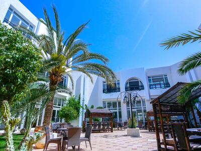 Innenhof mit weißen Gebäuden, Palmen, Tischen und Stühlen unter klarem blauem Himmel.