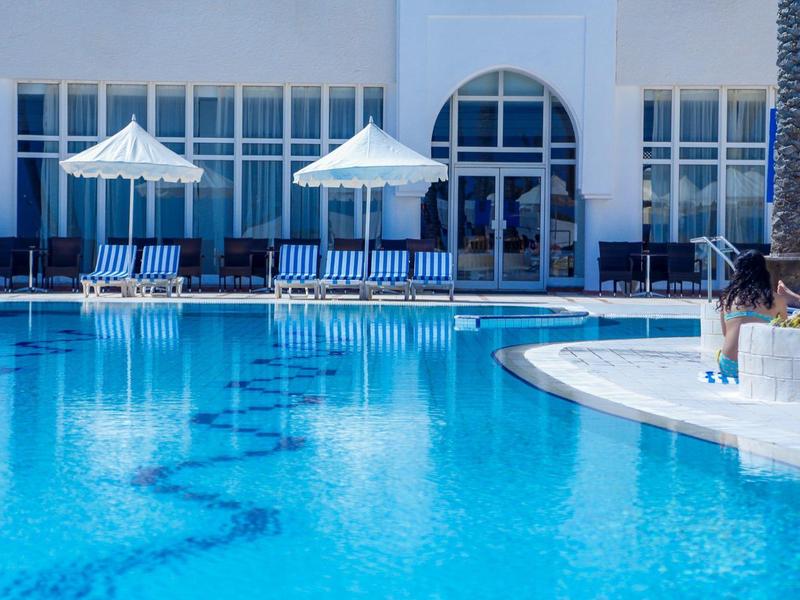 Großer blauer Pool mit Liegestühlen und weißen Sonnenschirmen vor einem weißen Gebäude.