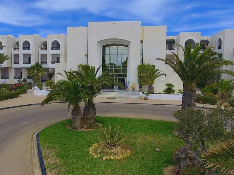 Weißes Gebäude mit Rundbögen, Palmen und grünem Vorgarten unter blauem Himmel.