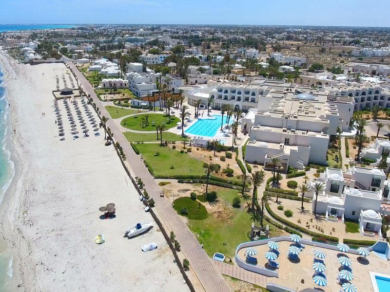 Langer Sandstrand mit klarem blauem Wasser, angrenzend an Hotelanlagen und Grünflächen.