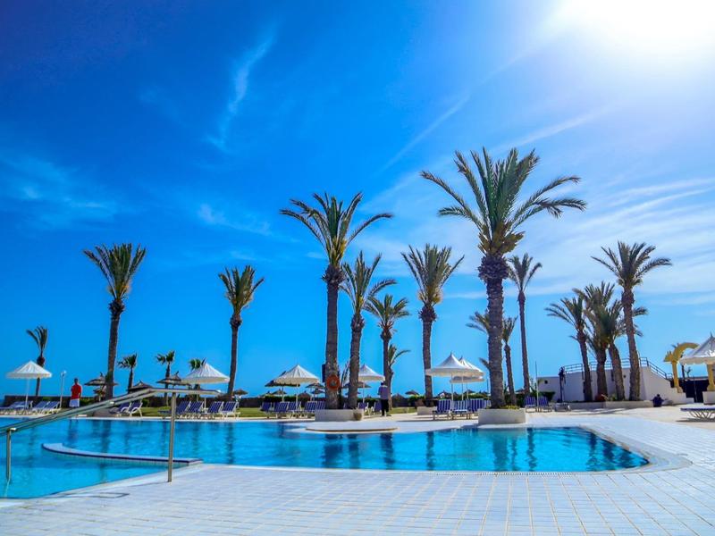 Ein klarer blauer Himmel über einem großen Pool mit Palmen und Sonnenschirmen in einer Resortanlage.