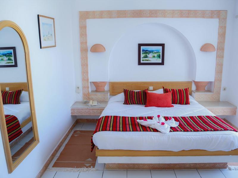 Ein helles Schlafzimmer mit großem Bett, bunten Kissen, Wandspiegel und zwei Nachttischen.