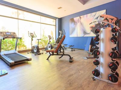 Heller Fitnessraum mit Hanteln, Laufband und anderen Trainingsgeräten vor großen Fenstern.