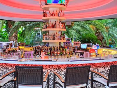 Moderne Bar mit rundem Tresen, bunten Flaschen, LED-Beleuchtung und leichten Stühlen vor Palmen.