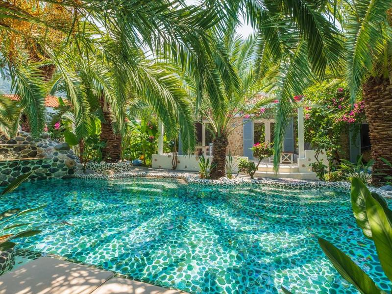 Blauer Pool mit Mosaikfliesen, umgeben von grünen Palmen vor einem hellen Haus.