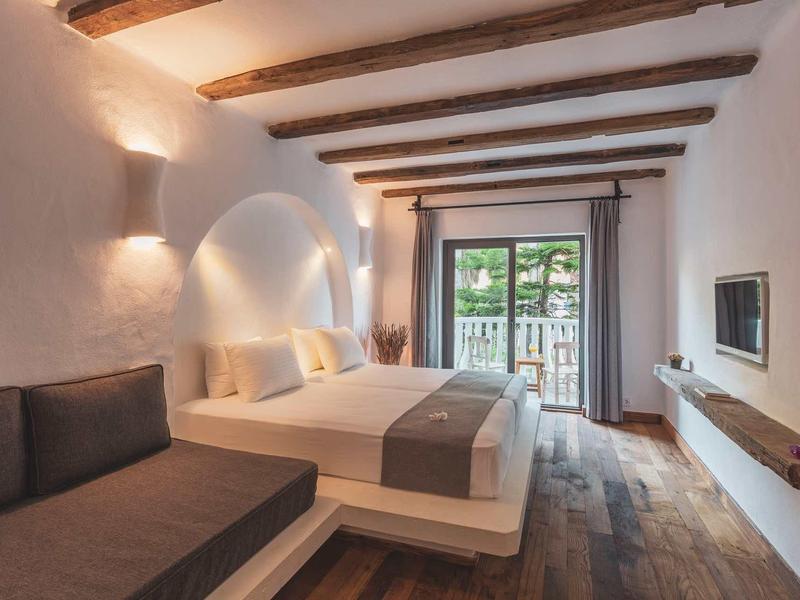 Helles Schlafzimmer mit Holzbalken, Doppelbett, Sofa, Holzboden und Balkon mit Grünblick.