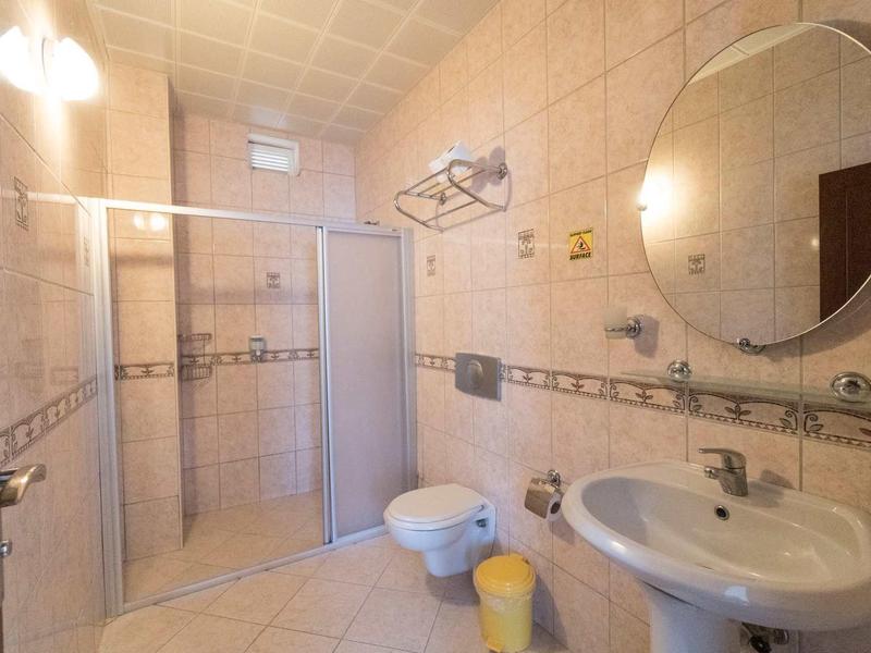 Světla koupelna se zaobleným zrcadlem, umyvadlem, toaletou a sprchovým koutem v béžových dlaždicích.