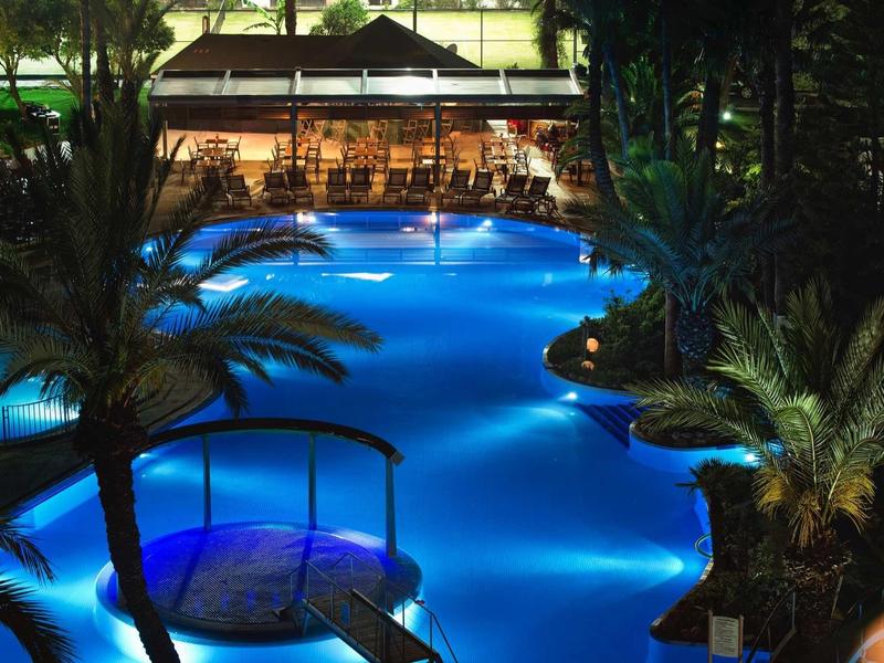 Piscina iluminada de noche rodeada de palmeras en un hotel tropical.