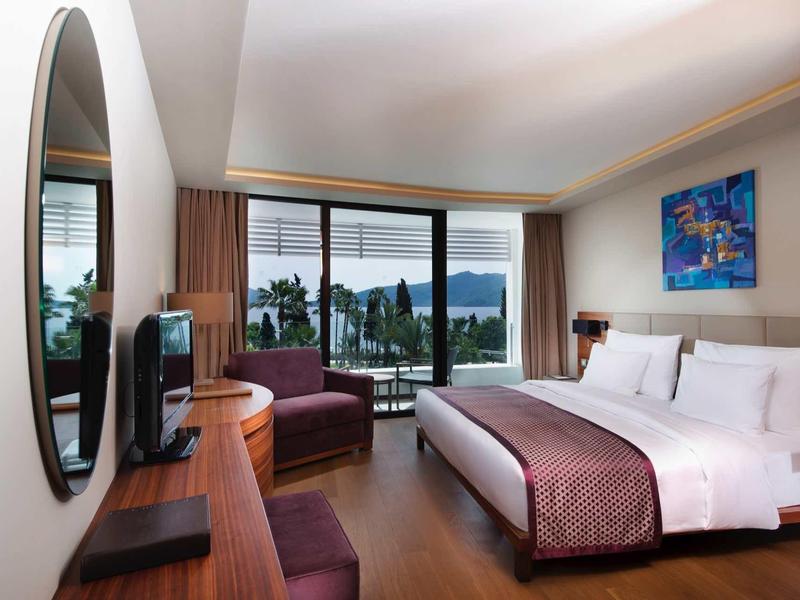 Habitación de hotel moderna con cama grande, zona de estar y vista a una terraza y al mar.