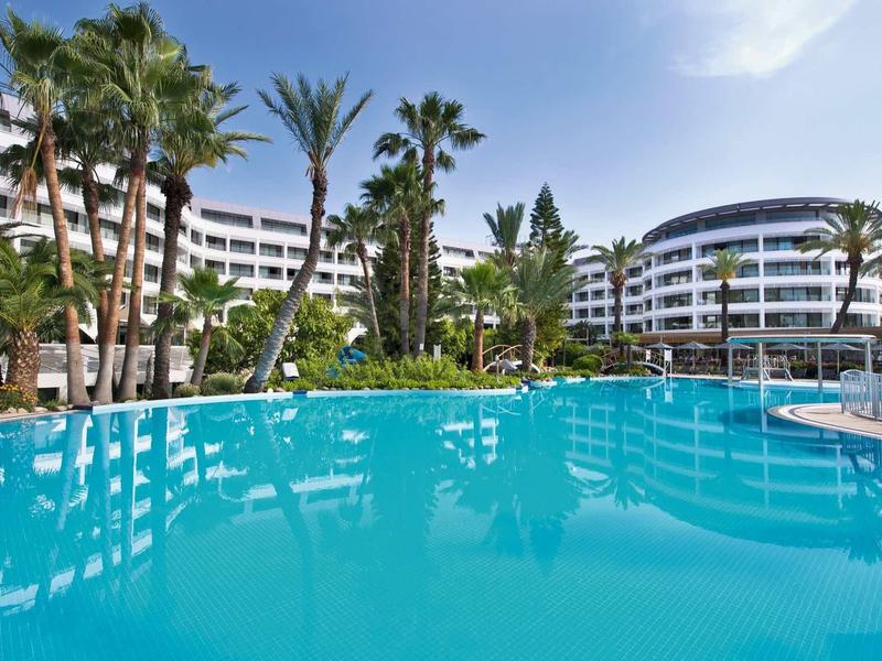 Gran piscina al aire libre frente a modernos edificios de hotel con muchas palmeras bajo cielo despejado.