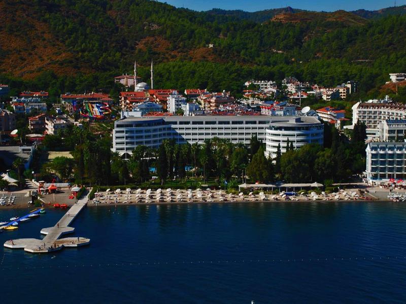 Gran hotel en la costa con tumbonas y muelle en el mar frente a colinas boscosas.