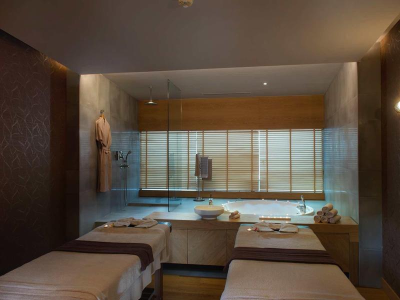 Sala de spa con dos camas de masaje, luz cálida y una ventana grande con cortinas en un hotel.