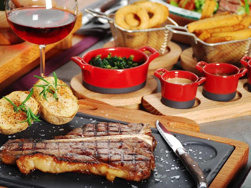 Parrilla con filete de carne, pan, guarniciones y copa de vino en una mesa de restaurante.