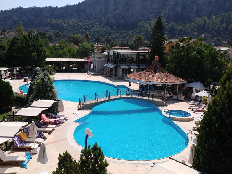 Leisure area with two blue swimming pools, sun loungers, and umbrellas surrounded by trees and mountains.