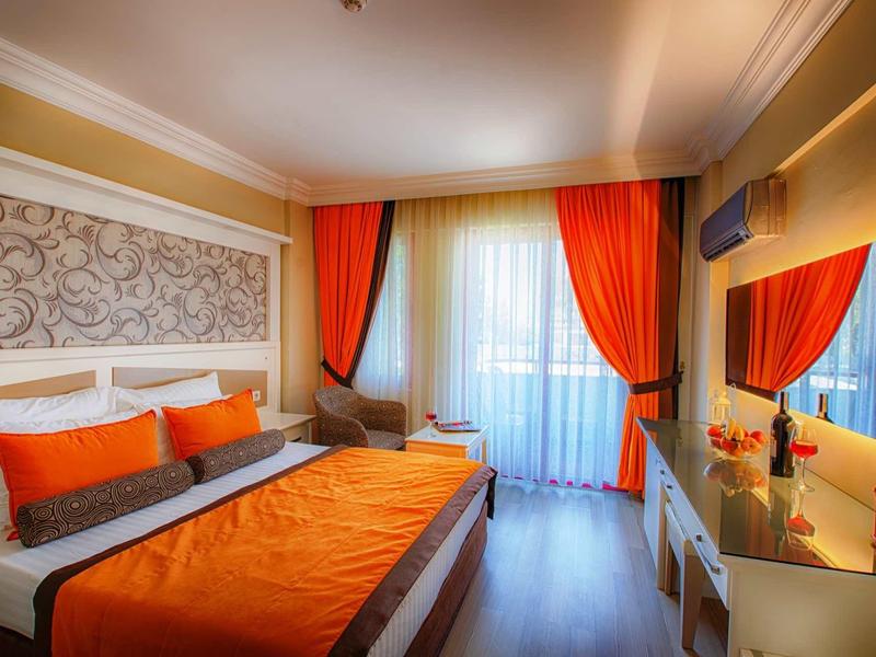 Chambre d'hôtel moderne avec lit double, literie et rideaux orange, sol en bois clair et balcon.