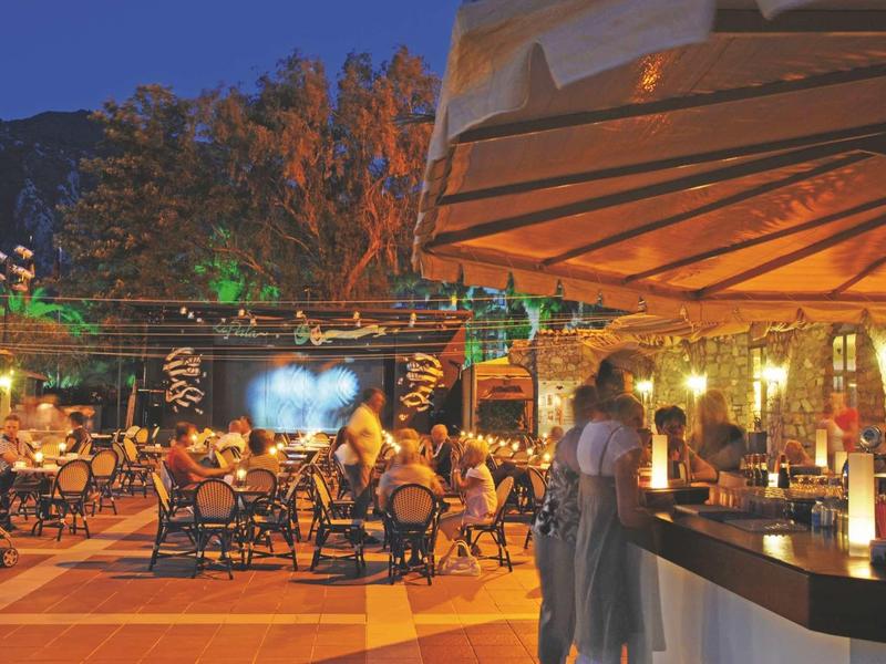 Eine Terrasse mit Tischen, Stühlen und Menschen bei Abendlicht unter einem Baum und einem Sonnenschirm.