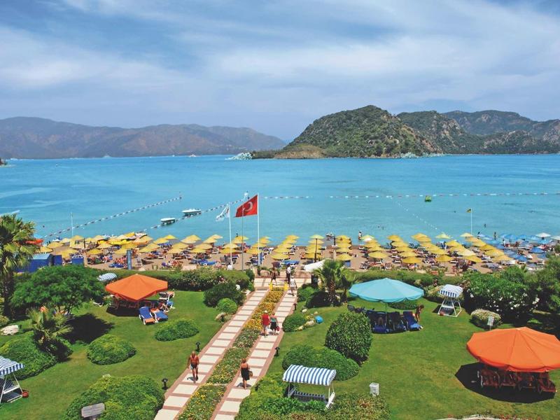 Strand mit bunten Sonnenschirmen, grüner Wiese und blauem Meer vor Inseln und Bergen.