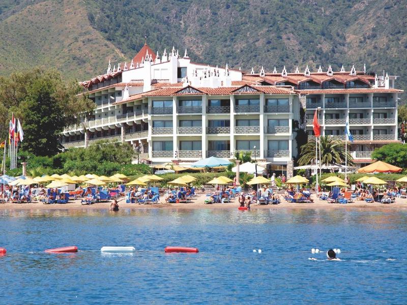 Hotel mit Glasfassade am Strand, davor Liegestühle, Sonnenschirme und blaues Meer.