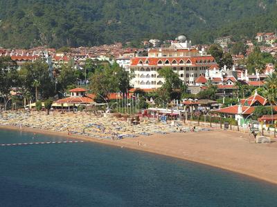 Strand mit Sand, Sonnenschirmen und Liegen vor einem Küstenort mit Gebäuden und bewaldetem Hügel.