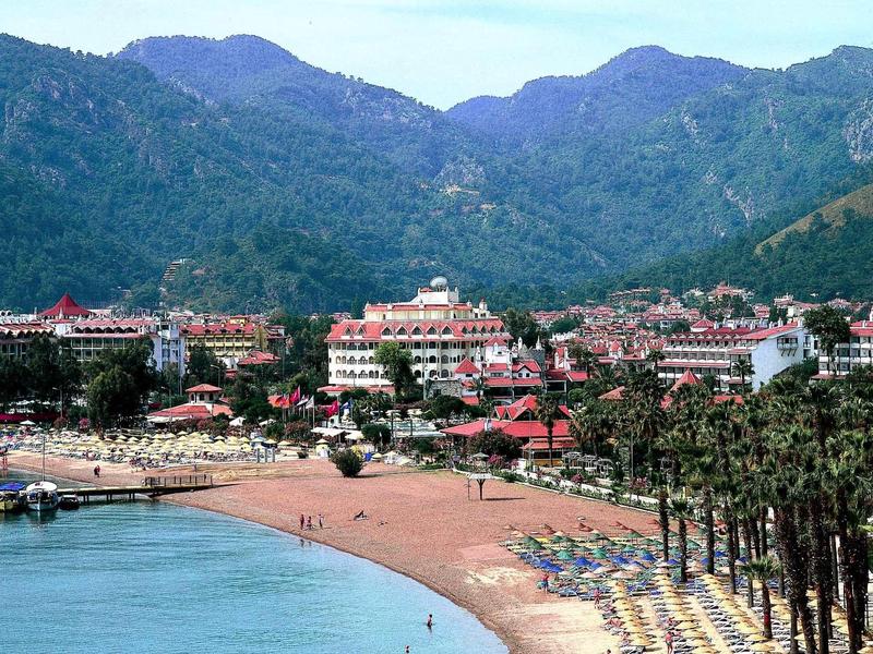 Küstenstadt mit Sandstrand, Palmen, Liegestühlen und bergen im Hintergrund unter blauem Himmel.