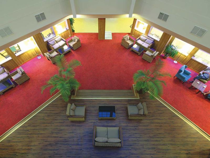 Großer Raum mit rotem Teppich, Sofas und Sesseln, hellgelben Wänden und Pflanzen.