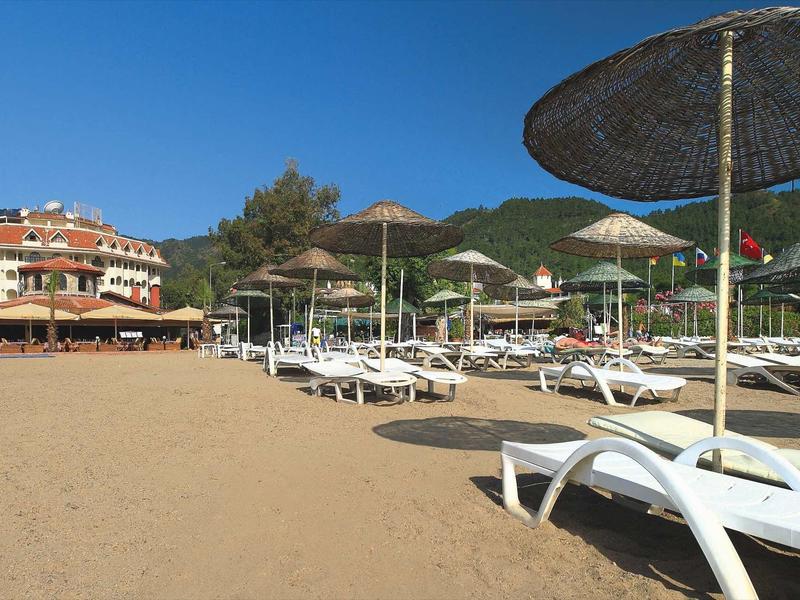 Sandstrand mit weißen Liegestühlen und Sonnenschirmen vor grünen Hügeln und blauen Himmel.