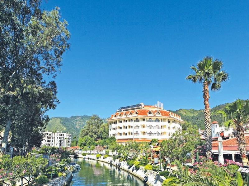 Fluss mit grünen Ufern, Palmen und einem großen runden Gebäude vor blauem Himmel.