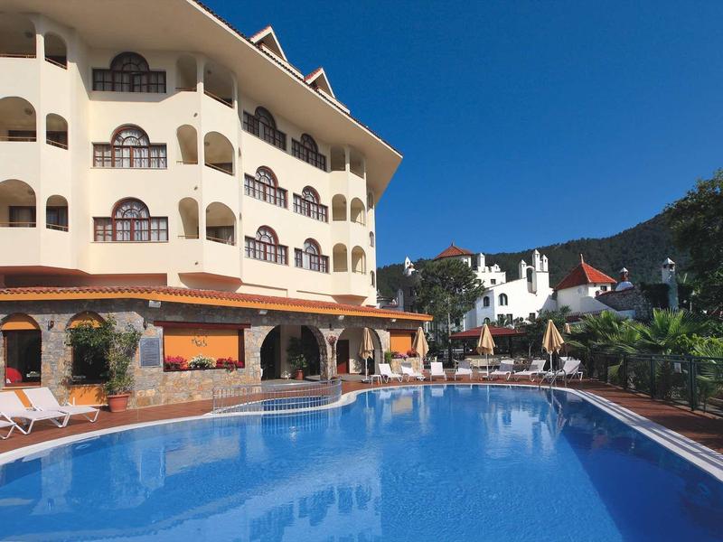 Großes Hotelgebäude mit Balkon, blauem Pool und Liegestühlen unter klarem Himmel.