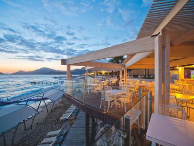 Strandrestaurant mit Holzterrasse, Glasgeländer und Blick auf Meer, Strand und Berg am Abend.