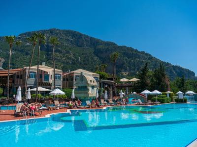 Großer Außenpool mit Sonnenschirmen und Liegestühlen vor einem Hotel und Bergen im Hintergrund.