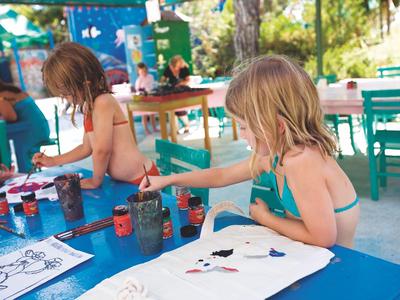 Kinder malen an einem Tisch im Freien mit Farbe und Pinseln, umgeben von bunten Stühlen.