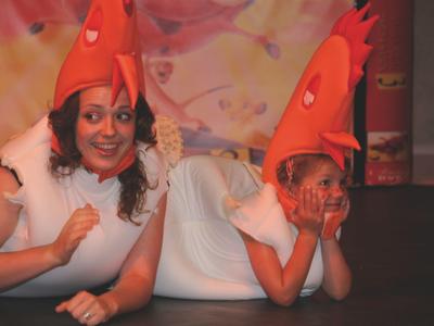 Zwei Personen in weißen Kleidern und roten Hühnerhüten liegen auf dem Boden und lächeln.