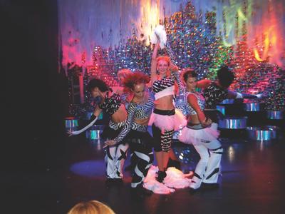 Drei in bunten Kostümen tanzende Künstler auf einer beleuchteten Bühne mit funkelndem Hintergrund.