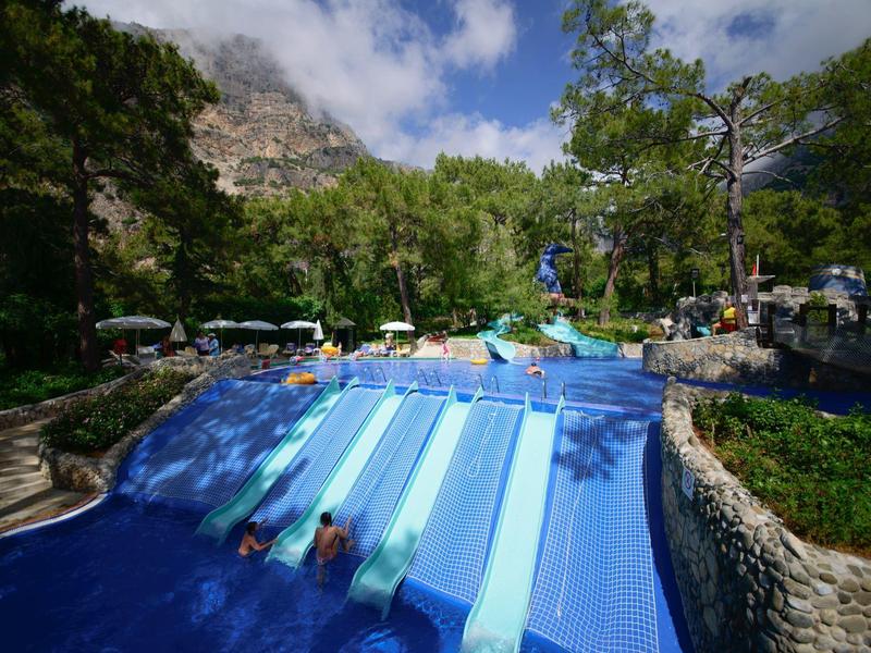 Mehrere Wasserrutschen in Blau, umgeben von Bäumen und Bergen bei sonnigem Himmel.