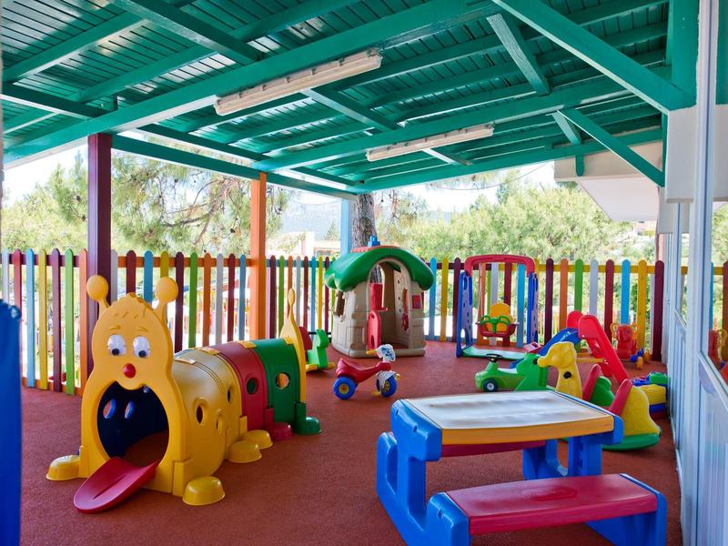 Überdachter Spielplatz mit bunten Spielgeräten und einem mehrfarbigen Holzzaun im Hintergrund.
