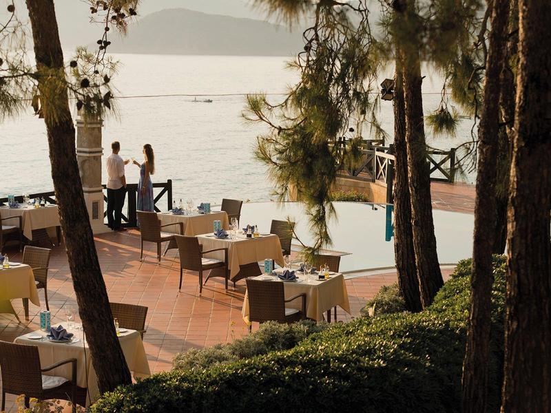 Terrasse mit Tischen und Stühlen am Meer, Bäume und Menschen beim Sonnenuntergang am Wasser.