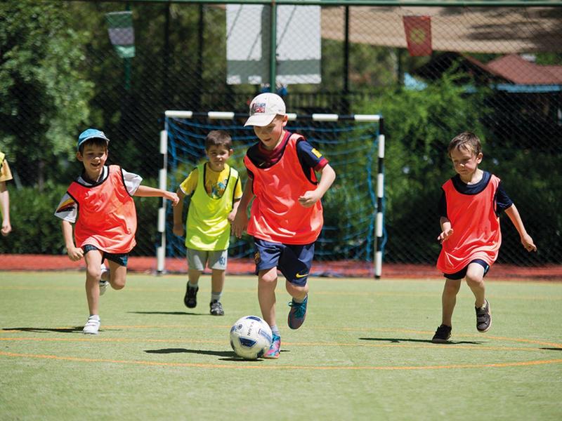 Vier Kinder rennen mit roten und gelben Westen auf einem Fußballfeld, ein Ball ist vorne.