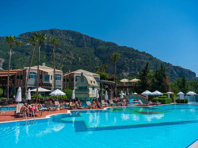 Großer, blauer Pool mit Liegestühlen und Sonnenschirmen vor bewaldetem Berg und klarem Himmel.