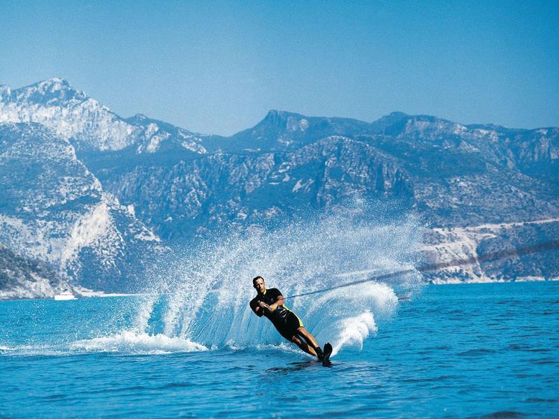 Ein Wasserskifahrer gleitet auf klarem Wasser vor schneebedeckten Bergen und klarem Himmel.