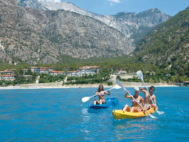 Vier Personen paddeln in zwei Kajaks auf blauem Wasser vor einer bergigen Küste mit Gebäuden.