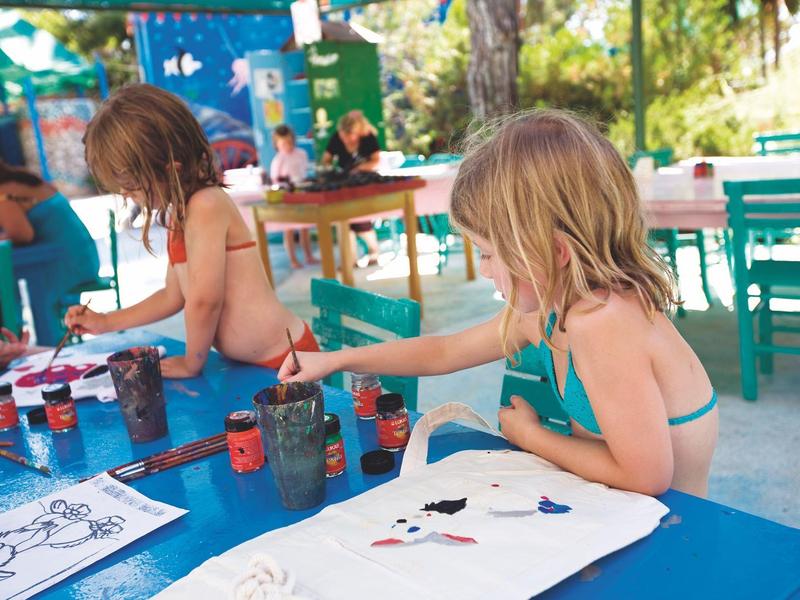 Kinder malen bunt auf Papier an einem sonnigen, draußen stehenden blauen Tisch.