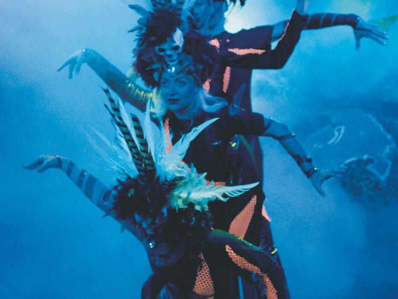 Drei Tänzerinnen mit Federkopfschmuck posieren vor blauem Hintergrund in elegantem Arrangement.