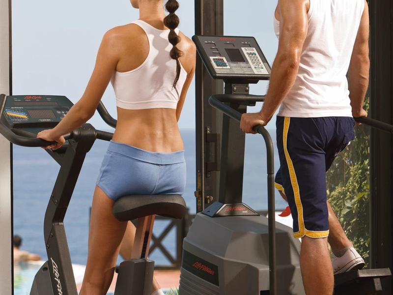 Zwei Personen trainieren auf Fahrradergometer und Stepper vor großen Fenstern mit Meerblick.