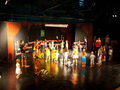 Kinder führen auf einer Bühne in einem Theaterstück mit bunten Kostümen und Bühnenbild auf.