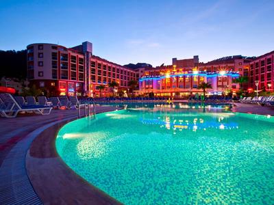 Large hotel with illuminated pool at night and lounge chairs by the poolside.