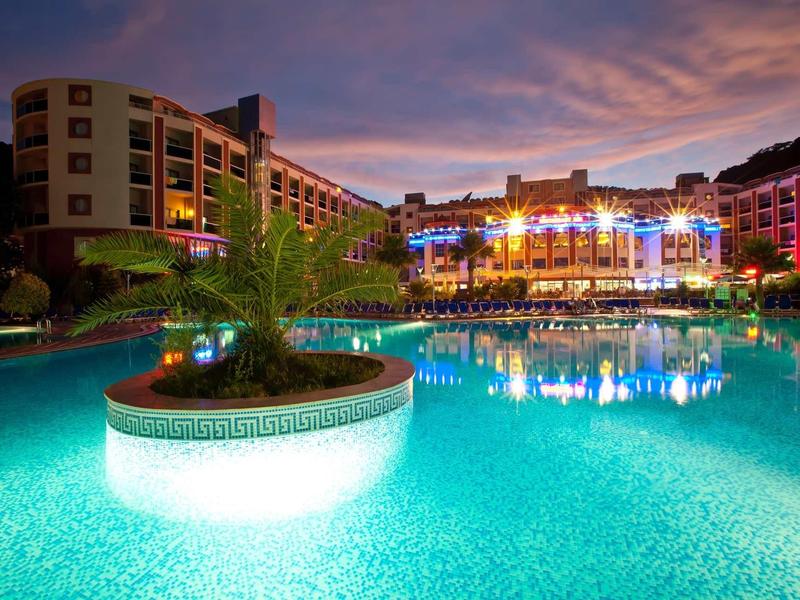 Illuminated outdoor pool with island and hotels in the background at sunset.
