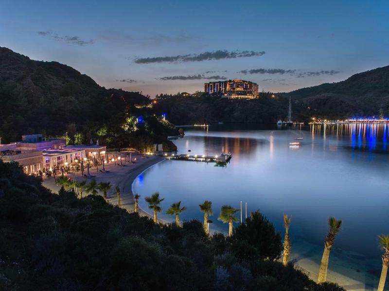 Osvětlená pláž za soumraku s kopci a hotelem v pozadí.